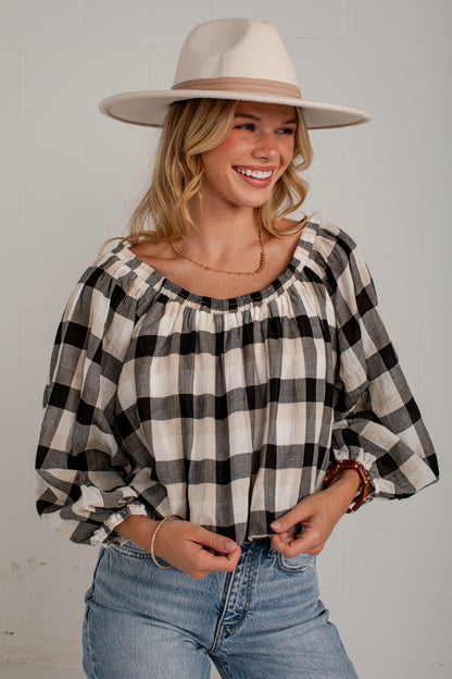 A woman wearing a black and white buffalo check plaid blouse with a wide scoop neckline and voluminous elbow-length sleeves, paired with light-wash blue jeans and a beige wide-brim hat, posing against a light gray brick wall.