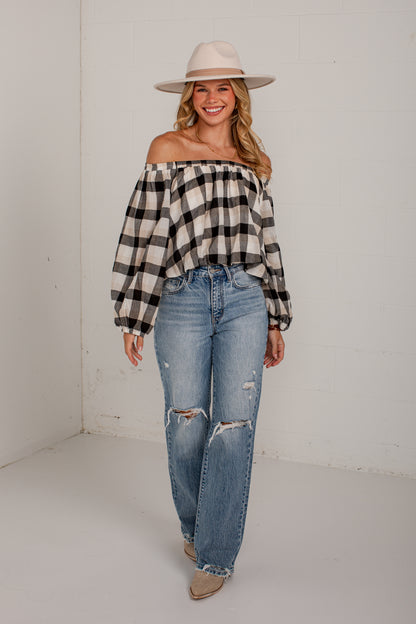 A woman modeling an off-the-shoulder black and beige buffalo plaid blouse with voluminous sleeves, paired with light-wash distressed blue jeans, beige boots, and a beige wide-brim hat, standing in a white studio corner.