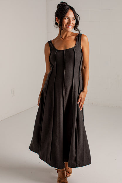 A woman modeling a sleeveless black midi dress with a structured bodice and flared skirt, standing in a bright white studio.