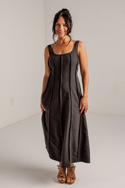 A woman modeling a sleeveless, midi-length black dress with vertical pleats, standing in a bright white studio.