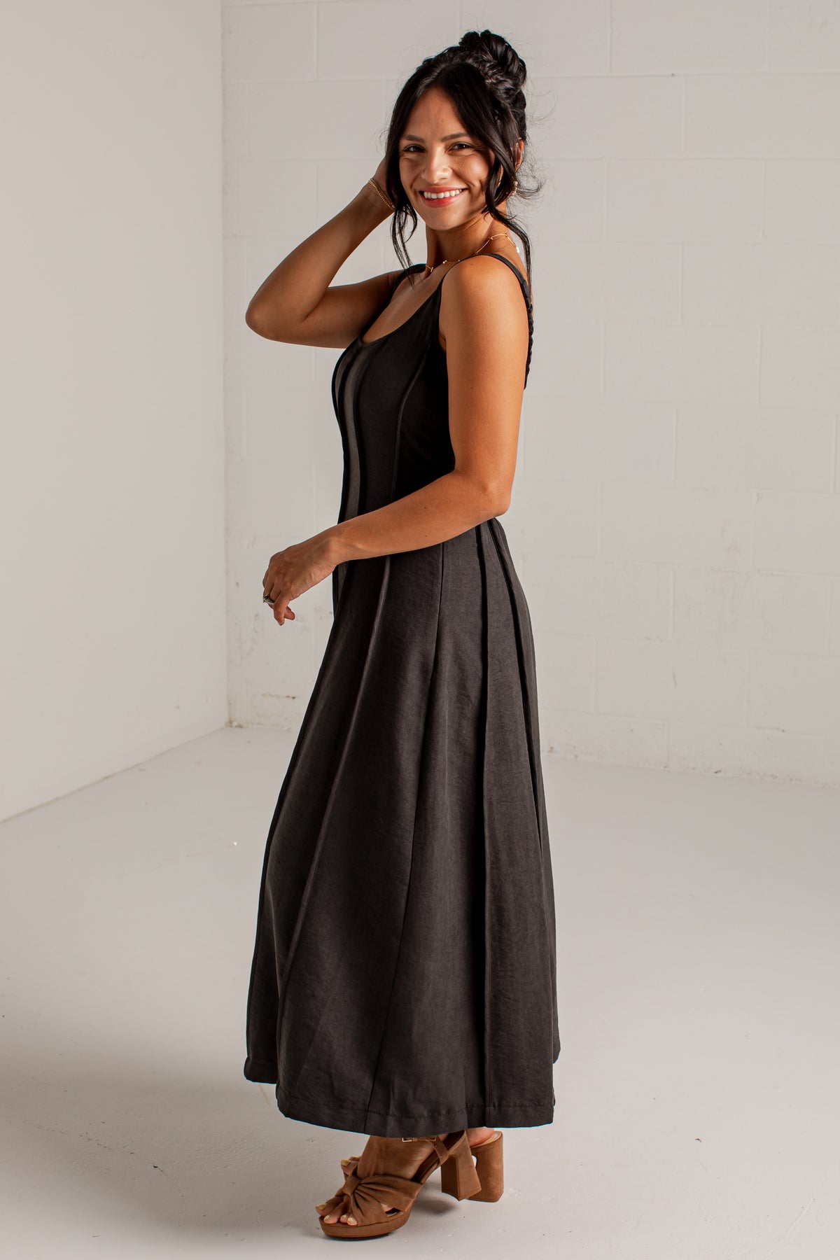 A woman modeling a sleeveless black midi dress with a pleated corset-style bodice and a flowing skirt, paired with brown heeled sandals, standing in a white studio corner.