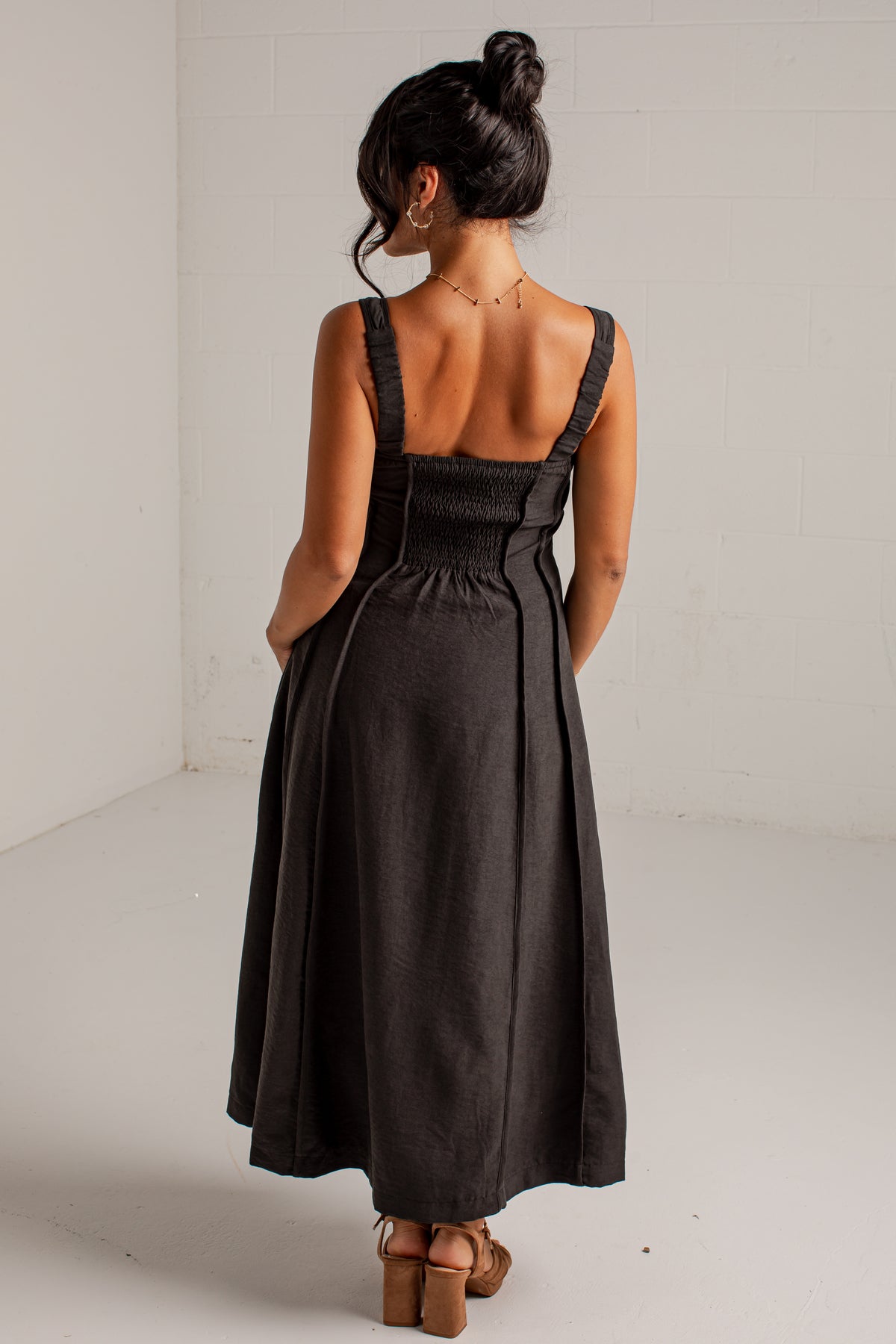 A woman viewed from behind wearing a dark charcoal gray midi dress with shoulder straps, a smocked bodice, and a flared skirt, paired with brown block-heel sandals in a bright studio setting.