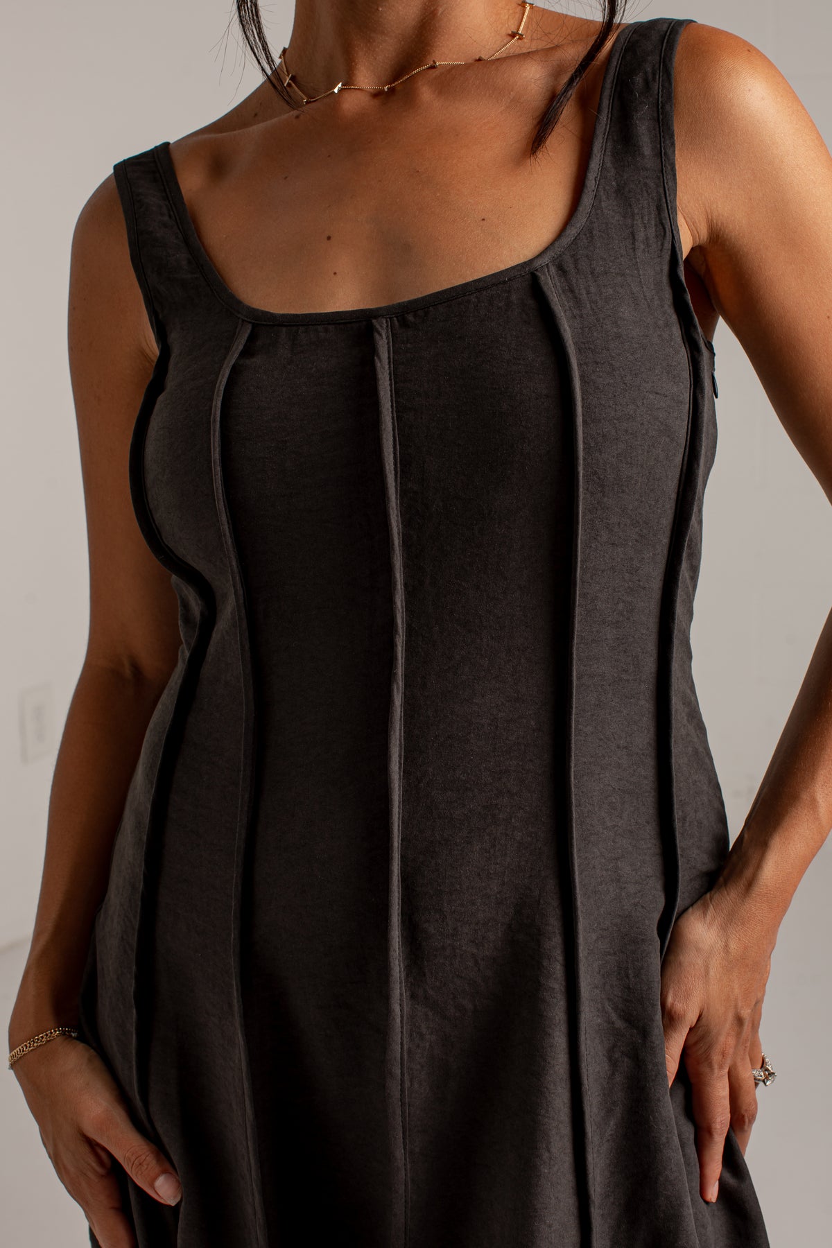 A close-up of a woman wearing a dark charcoal gray sleeveless dress with structured vertical seams and a square neckline.