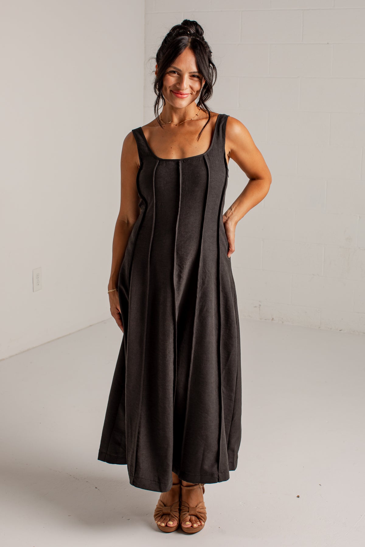 A woman modeling a sleeveless black maxi dress with a square neckline and vertical seam details, standing in a white studio setting.