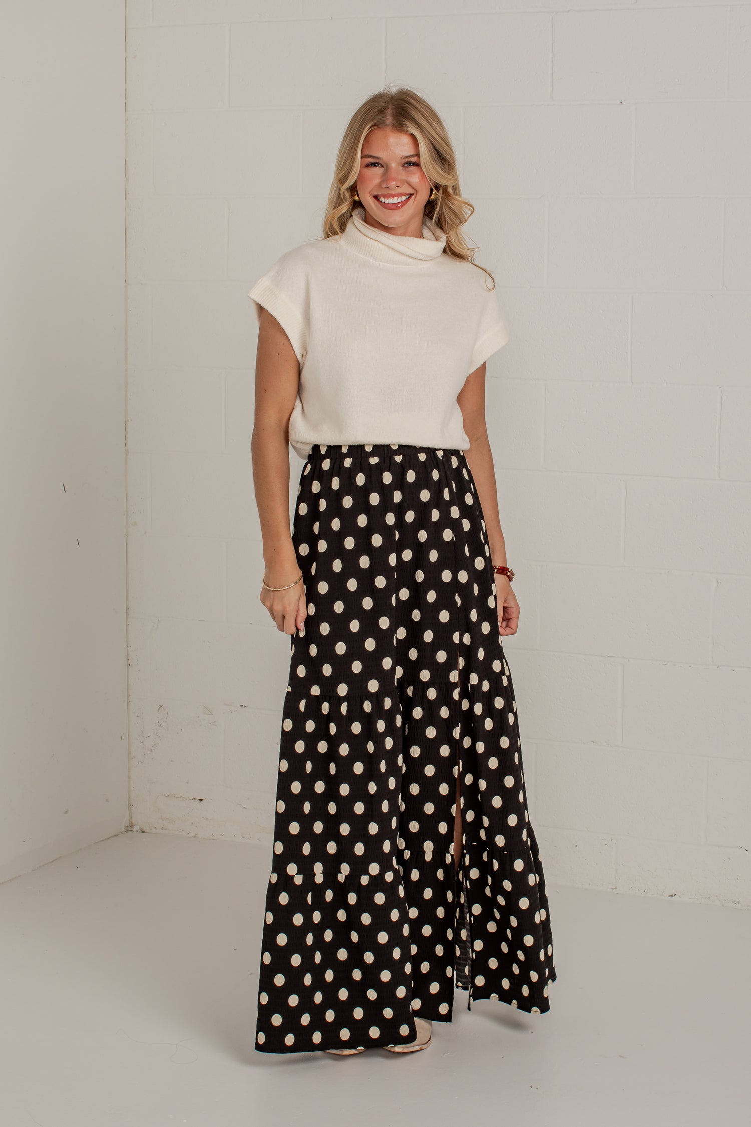 A woman modeling a cream short-sleeve mock neck top and a black maxi skirt with beige polka dots, standing in a white studio corner.