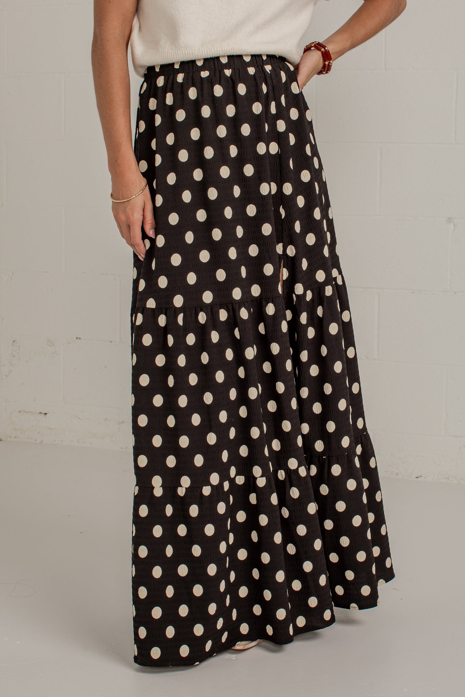 A woman wearing a black maxi skirt with a cream polka dot pattern, paired with a white top, standing against a white brick wall.