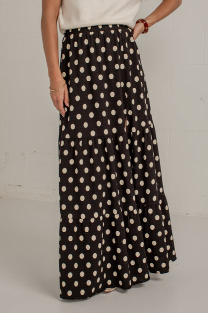 A woman wearing a black maxi skirt with a cream polka dot pattern, paired with a white top, standing against a white brick wall.
