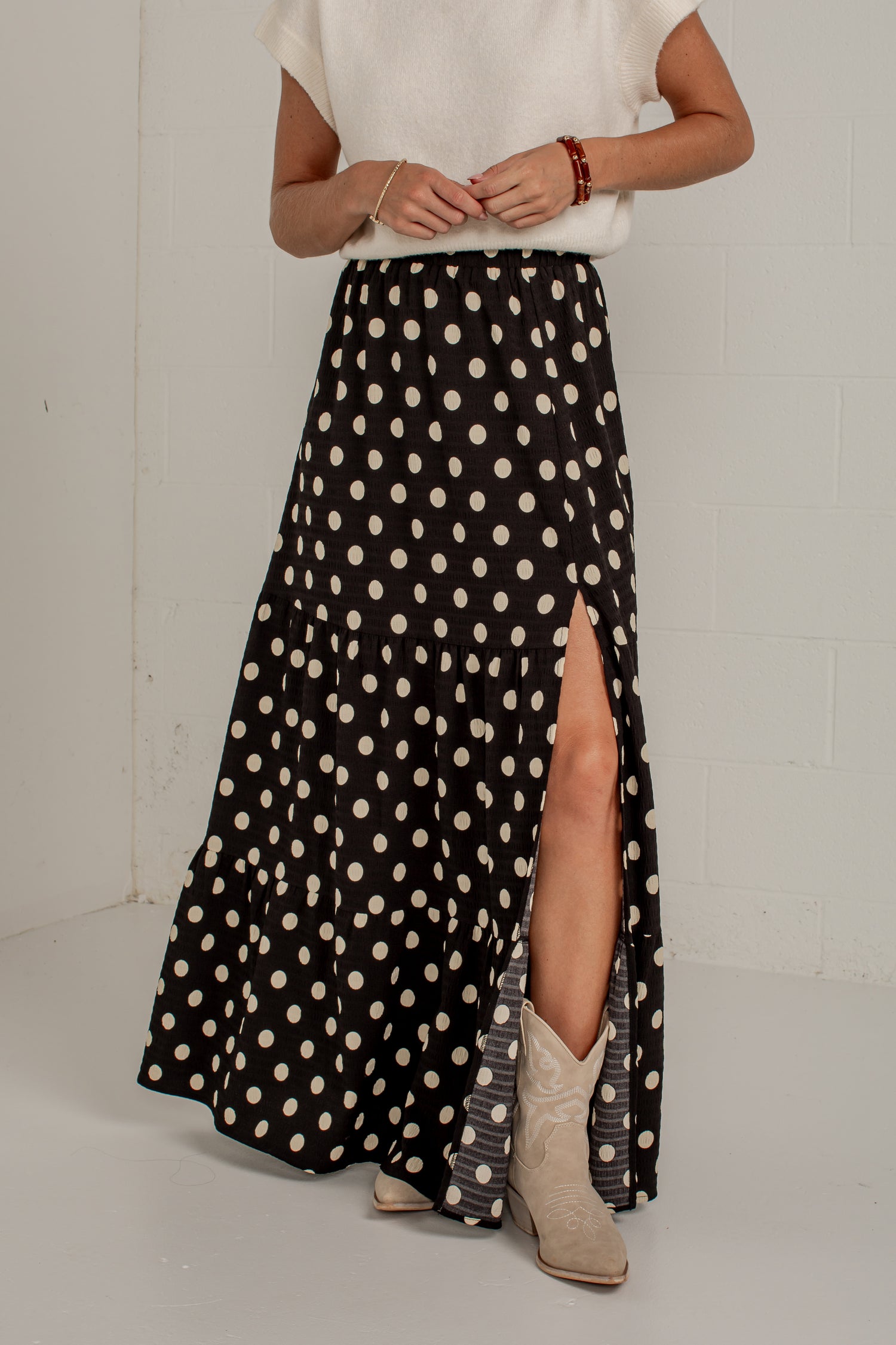 A woman wearing a long black skirt with beige polka dots and a high front slit, paired with a white top and beige cowboy boots, standing against a white brick wall.