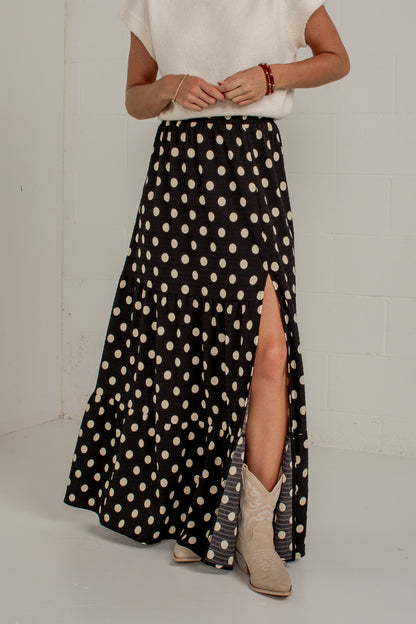 A woman wearing a long black skirt with beige polka dots and a high front slit, paired with a white top and beige cowboy boots, standing against a white brick wall.