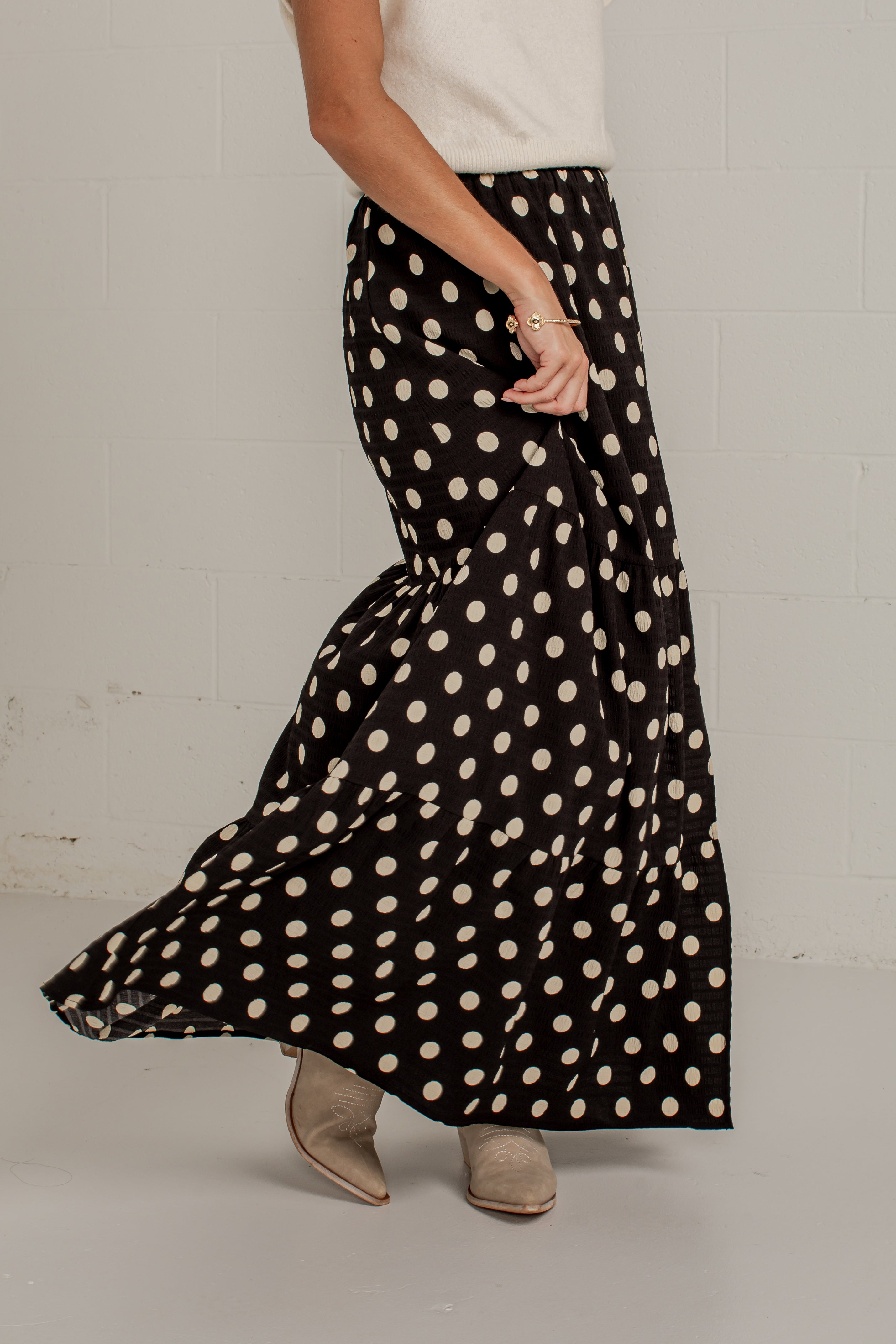A person wearing a long black skirt with large cream polka dots, paired with a white top and beige ankle boots, standing against a light gray wall.