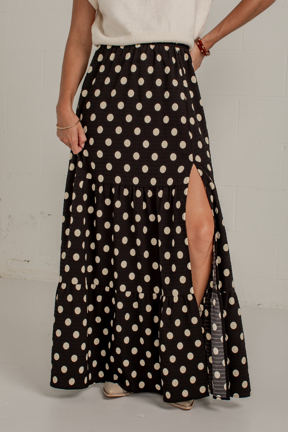 A woman wearing a long black maxi skirt with a cream polka dot pattern and a high side slit, paired with a cream top against a light gray wall.
