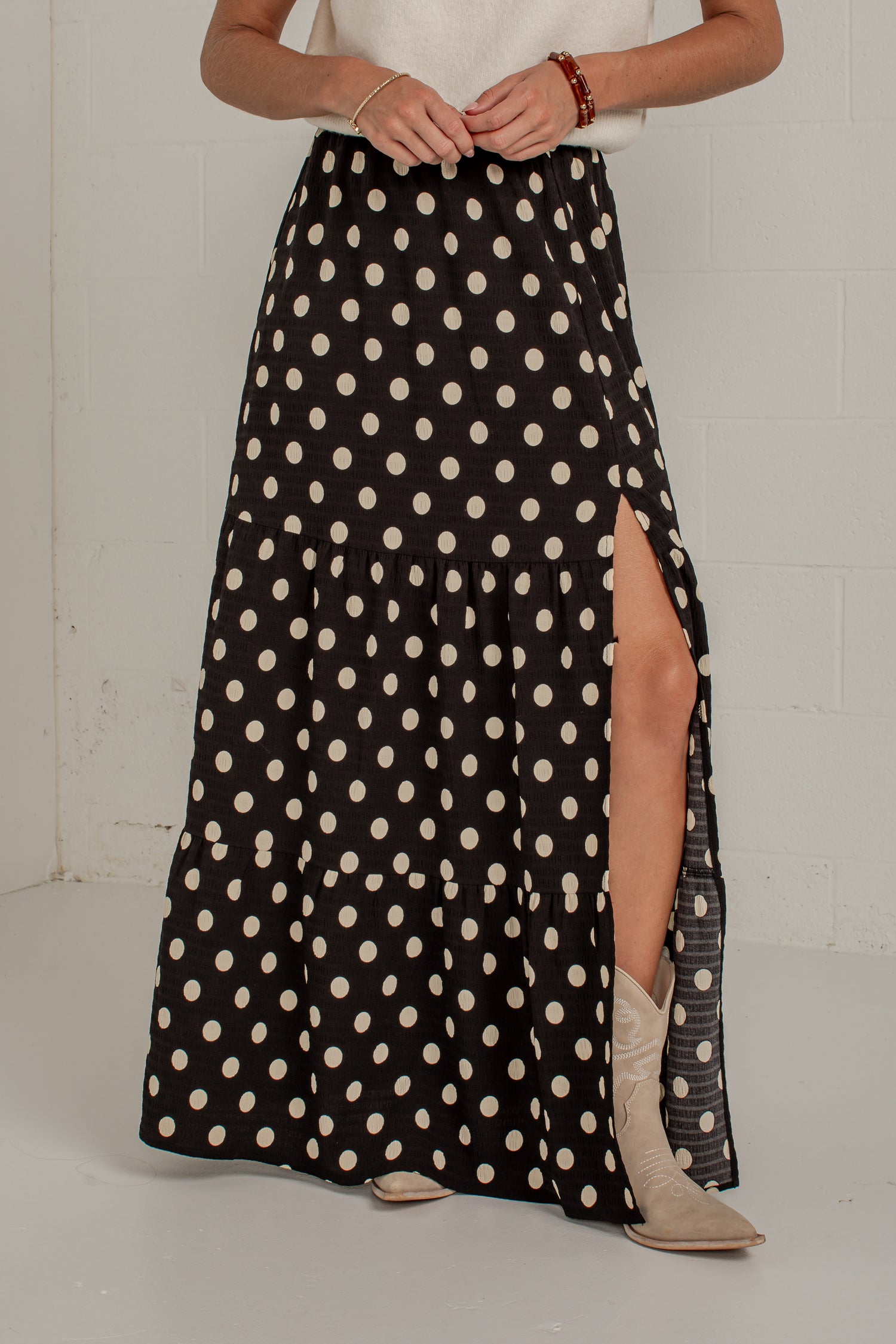 A black maxi skirt with a cream polka dot pattern and a high side slit, worn by a model in a white top and beige boots against a white brick wall.