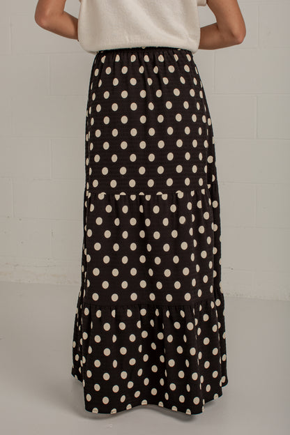 A black tiered maxi skirt with a cream polka dot pattern, shown on a model from the back against a light gray brick wall.