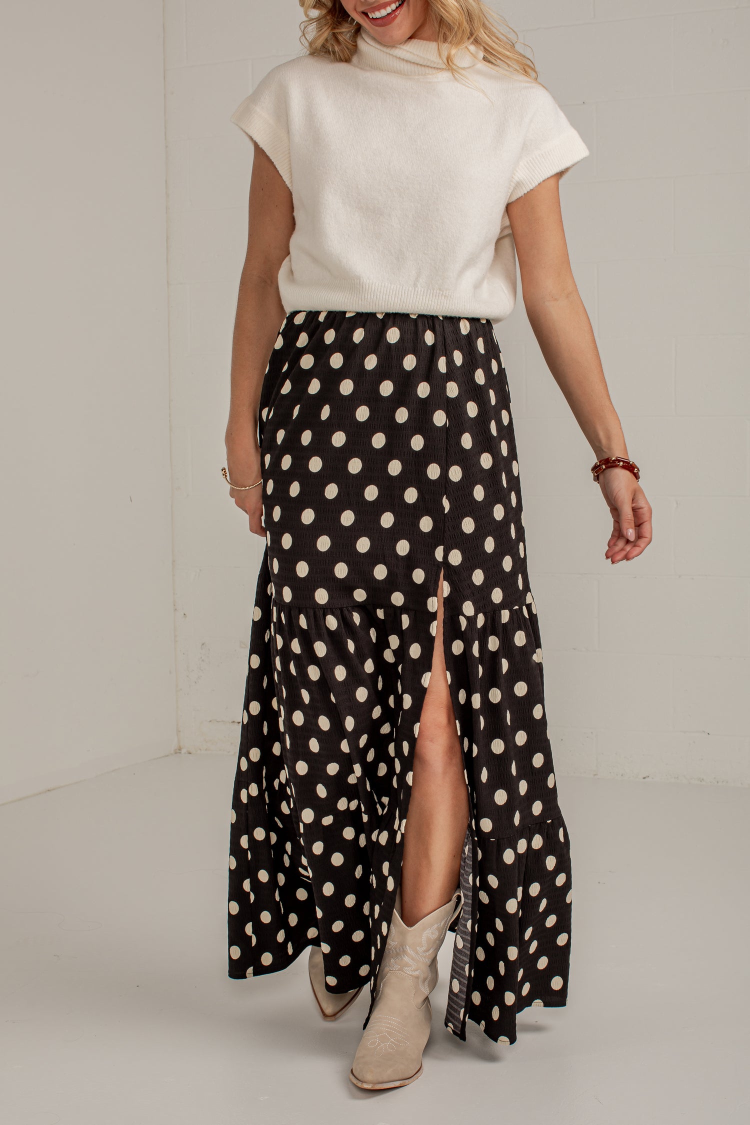 A woman wearing a black midi skirt with a cream polka dot pattern and a front slit, styled with a cream knit top and beige ankle boots.