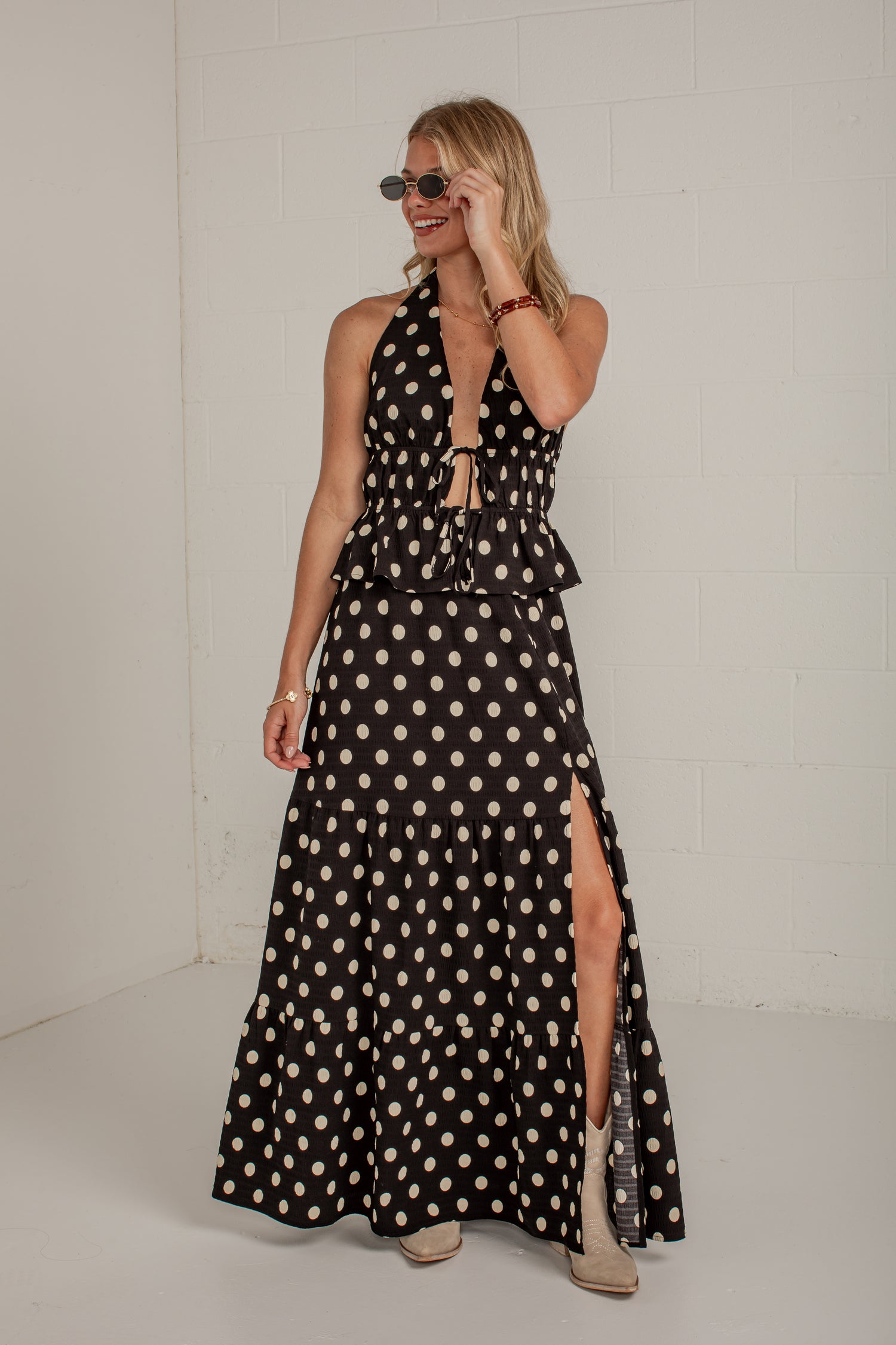 A woman modeling a sleeveless black halter maxi dress with a beige and white polka dot pattern, featuring a tie-front waist and a high side slit, set against a white brick wall.