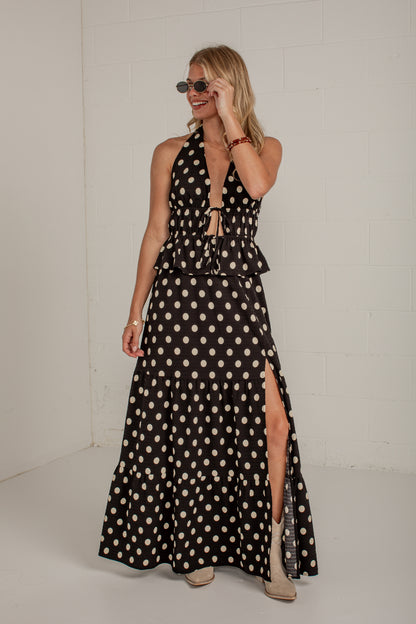 A woman modeling a sleeveless black halter maxi dress with a beige and white polka dot pattern, featuring a tie-front waist and a high side slit, set against a white brick wall.