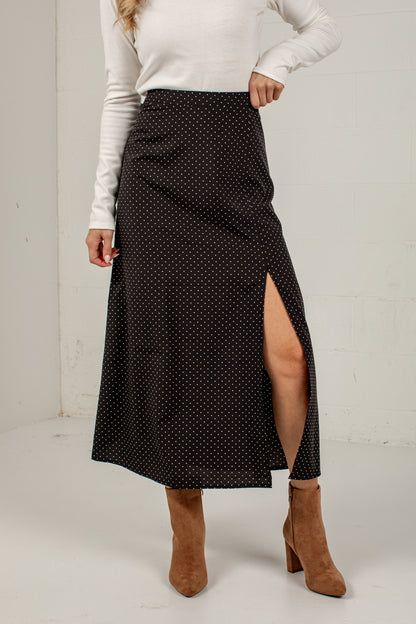 A woman models a black midi skirt with a small white polka dot pattern and a high side slit, paired with a white long-sleeve top and brown ankle boots.