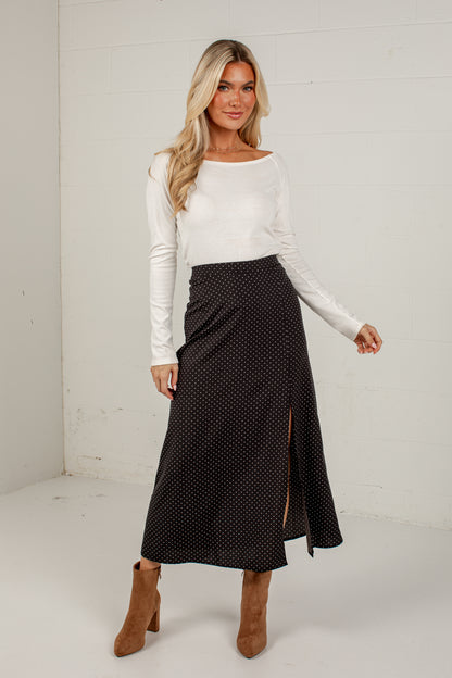 A woman modeling a black midi skirt with small white polka dots and a side slit, paired with a white long-sleeve top and brown ankle boots, standing against a white paneled wall.