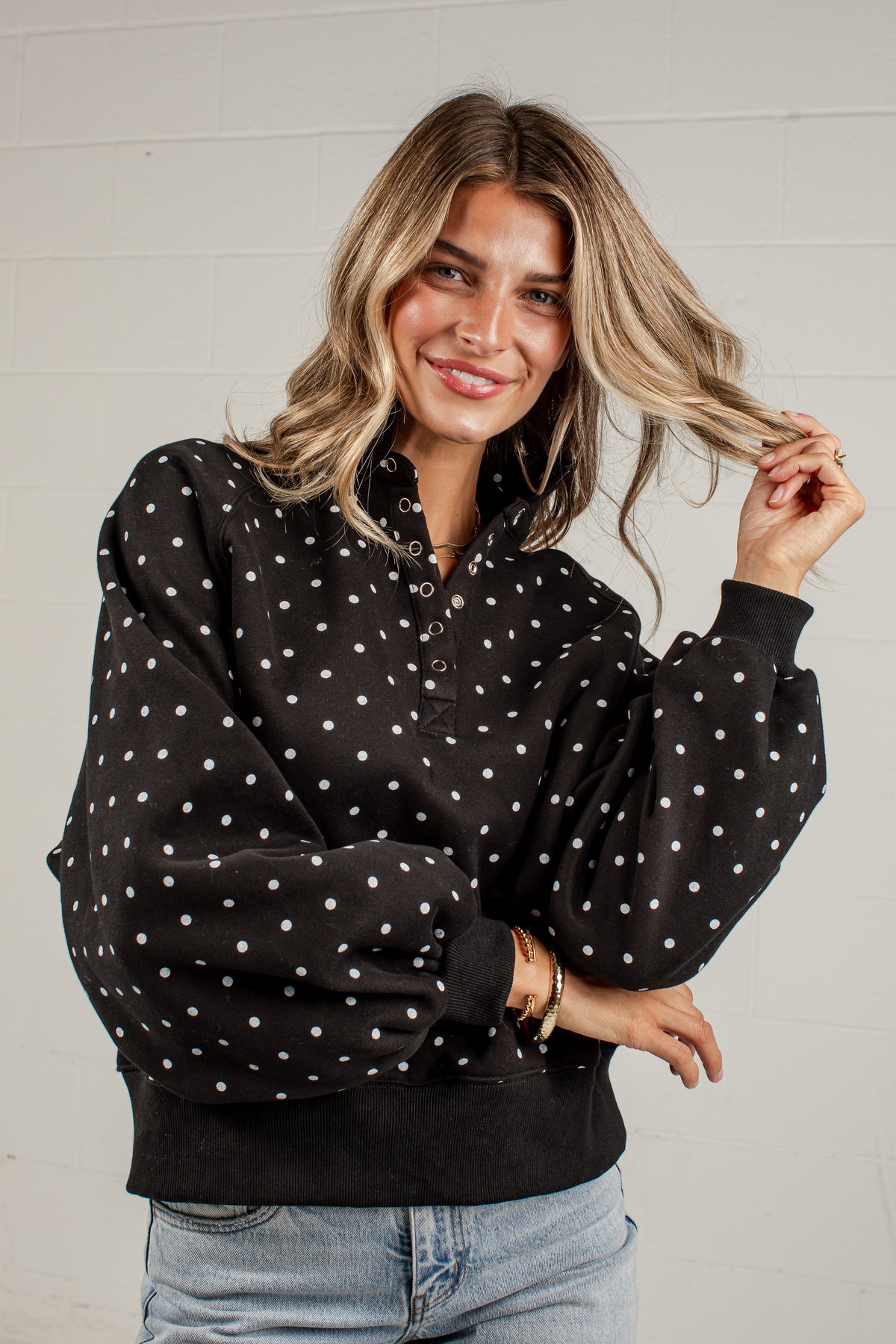 A woman wearing a black long-sleeve button-up top with white polka dots and voluminous balloon sleeves, paired with light blue jeans, posing against a white brick wall.