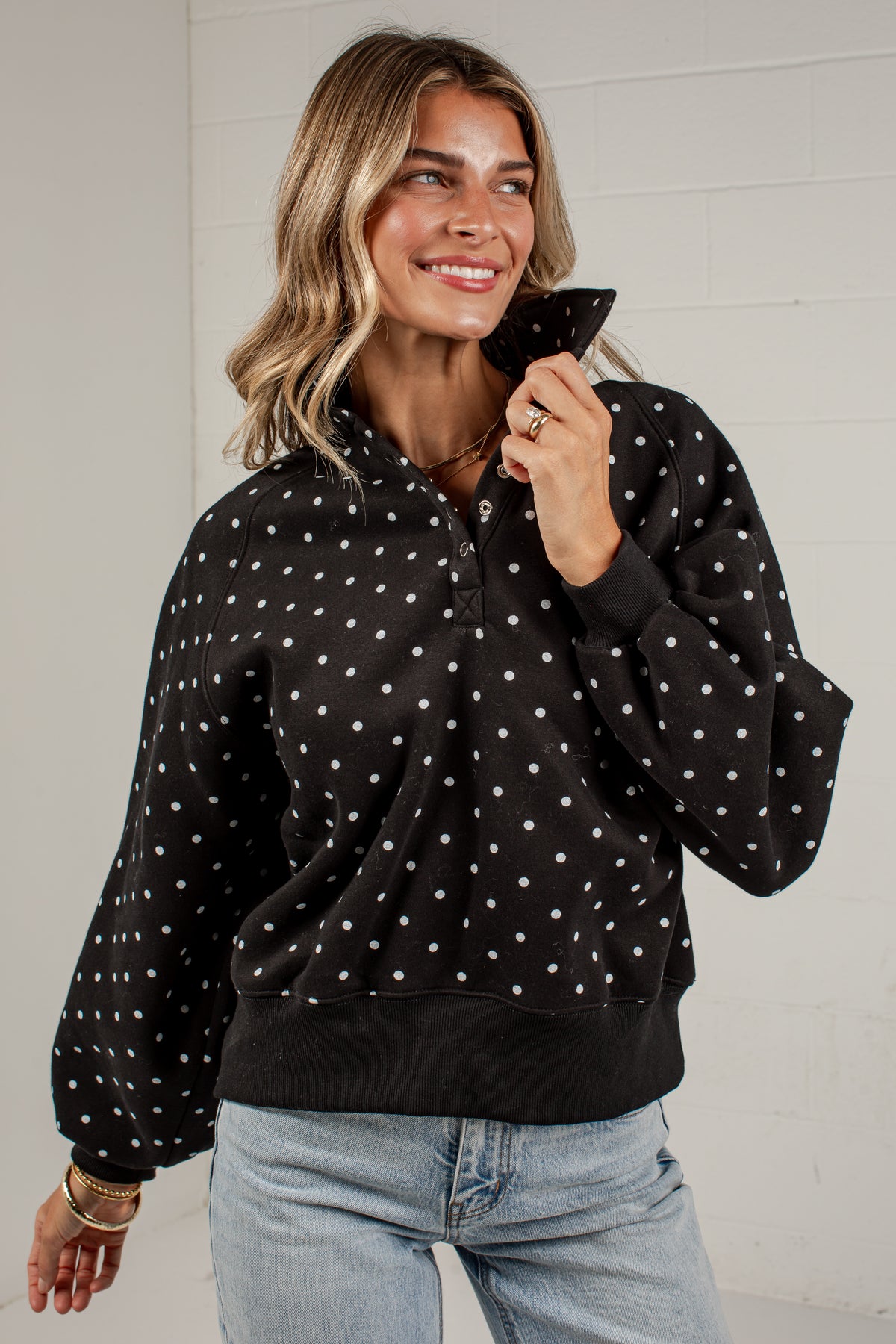 A woman wearing a black long-sleeve half-zip pullover sweatshirt with white polka dots, paired with light-wash blue jeans, standing against a white brick wall.
