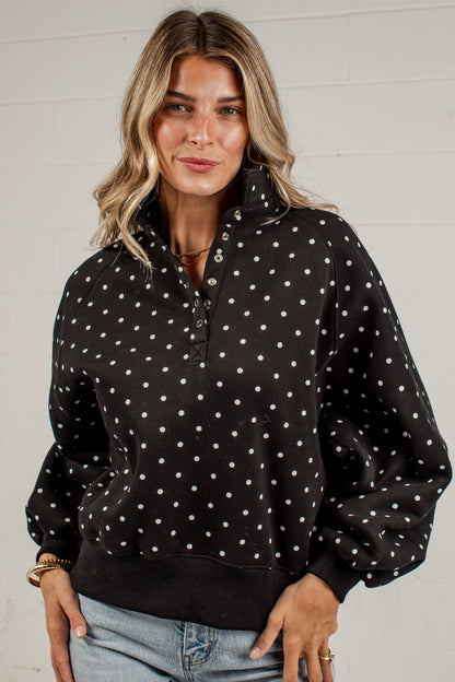 A woman wearing a black long-sleeve quarter-zip sweatshirt with white polka dots, paired with light-wash blue jeans, standing against a white brick wall.
