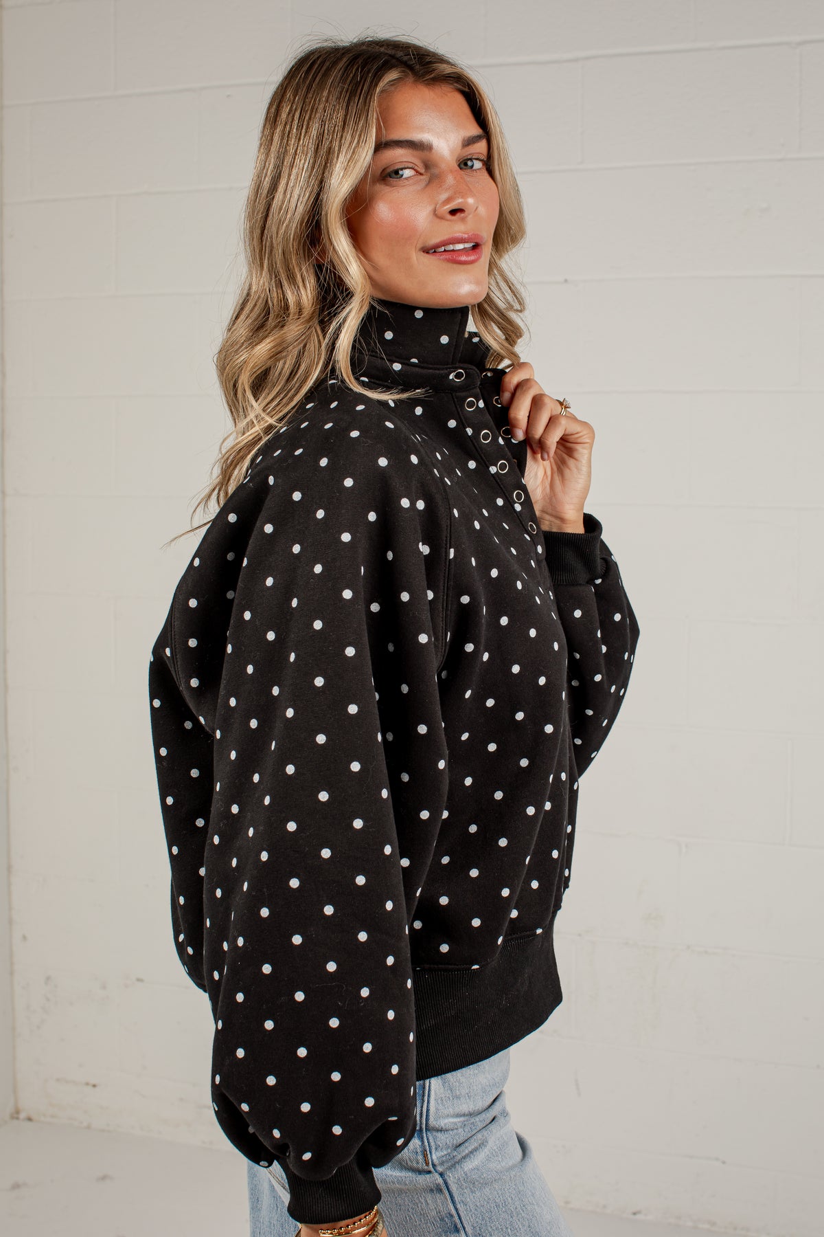 A woman wearing a black turtleneck sweatshirt with white polka dots, paired with light blue jeans, standing against a white brick wall.