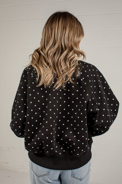 A woman viewed from behind wearing a black crewneck sweatshirt with white polka dots, paired with light blue jeans, standing against a white brick wall.