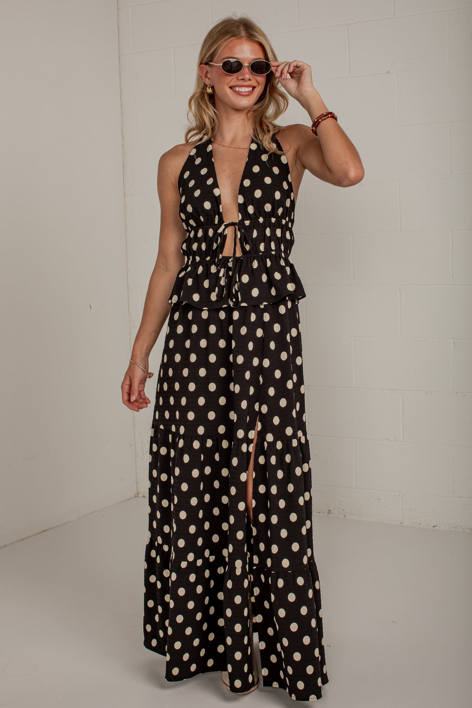A woman modeling a black halter-neck maxi dress with a beige polka dot pattern, featuring a deep V-neckline, tie details at the waist, and a thigh-high front slit.