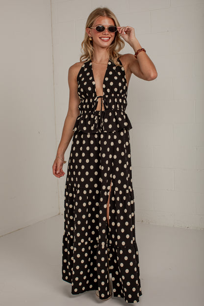A woman modeling a black halter-neck maxi dress with a beige polka dot pattern, featuring a deep V-neckline, tie details at the waist, and a thigh-high front slit.