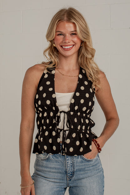 A smiling blonde woman wearing a black halter-style top with beige polka dots, a white camisole underneath, and light blue jeans, standing against a white brick wall.