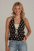A smiling blonde woman wearing a black halter-style top with beige polka dots, a white camisole underneath, and light blue jeans, standing against a white brick wall.