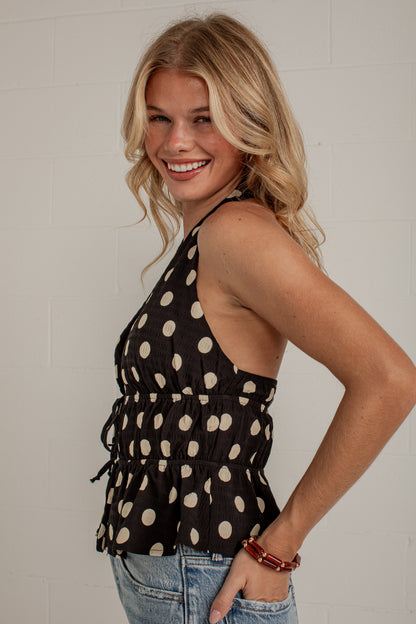 A smiling blonde woman wearing a black halter-style top with beige polka dots and blue jeans, standing against a light gray brick wall.