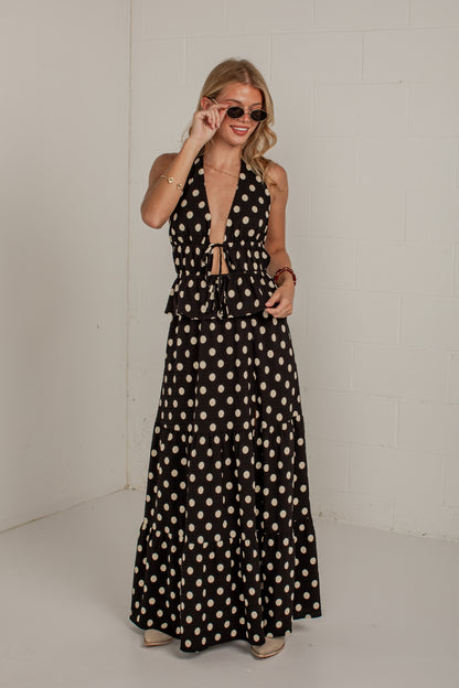 A woman modeling a black halter-neck maxi dress with a beige polka dot pattern, standing against a white brick wall.