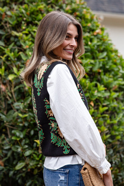 Woman wearing a black embroidered vest over a white blouse with blue jeans, standing in front of green bushes.