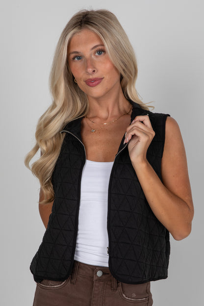 Woman wearing a black quilted vest over a white top and brown pants on a gray background