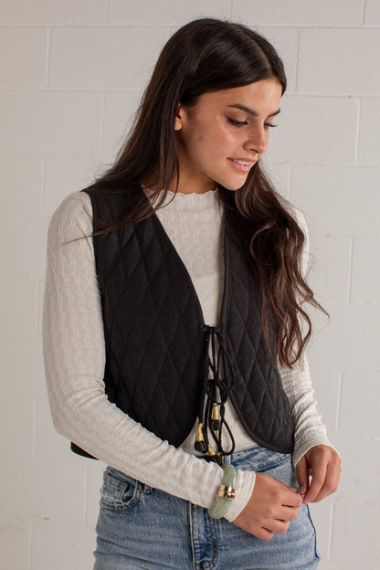 A woman wearing a black diamond-quilted sleeveless vest over a light gray ribbed long-sleeve top and blue jeans, standing against a white brick wall.