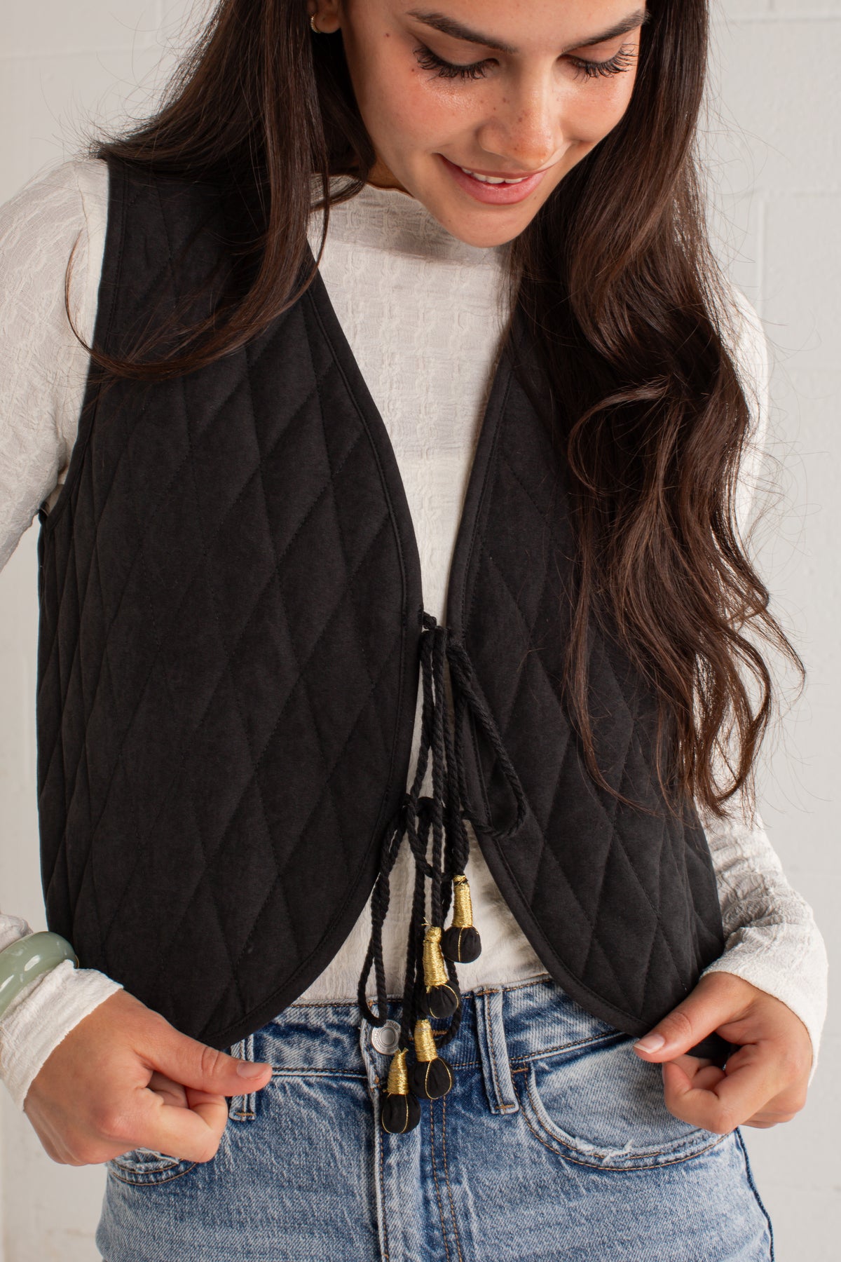 A woman wearing a black diamond-quilted sleeveless vest over a white long-sleeve top and blue jeans, with the vest tied at the front with black cords and gold-toned beads.