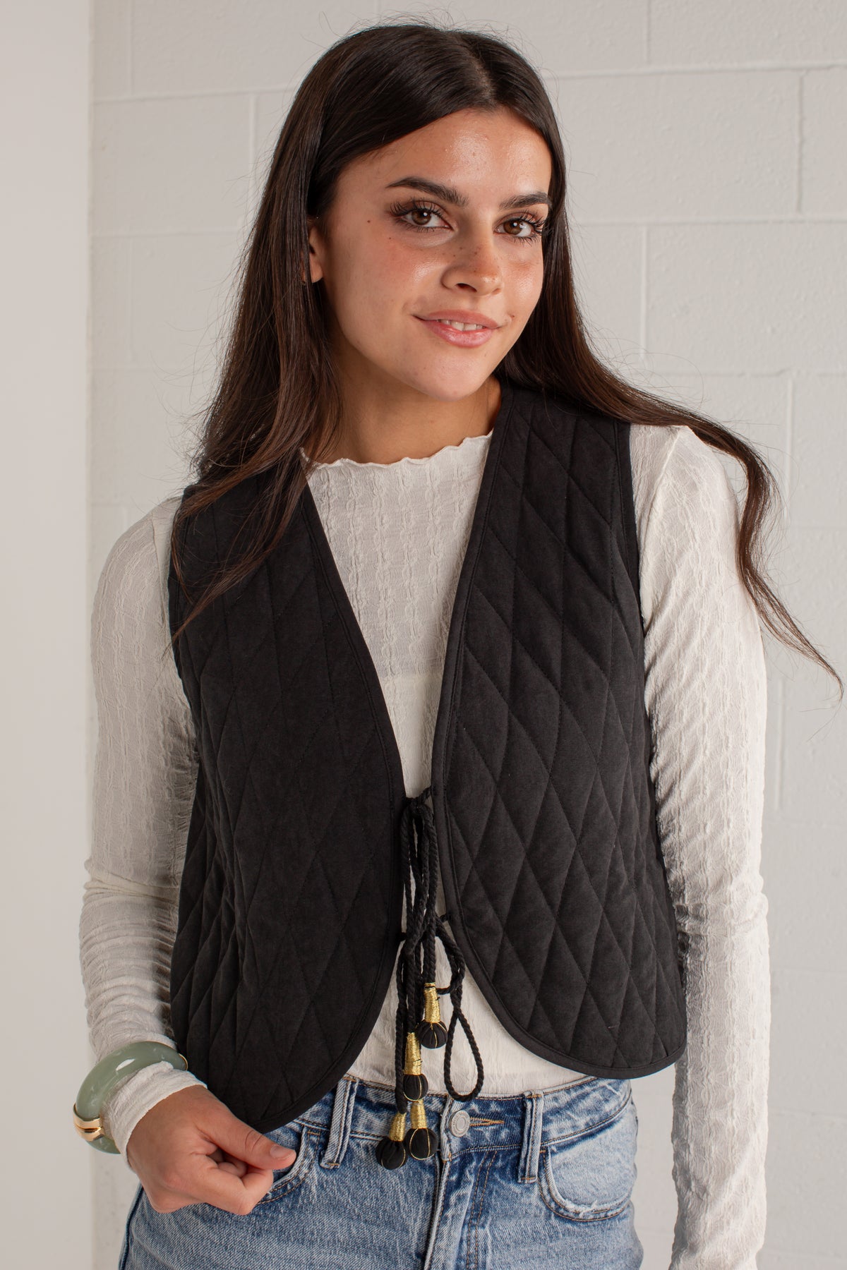 A woman wearing a black diamond-quilted sleeveless vest over a white ribbed long-sleeve top and blue jeans, standing against a white brick wall.