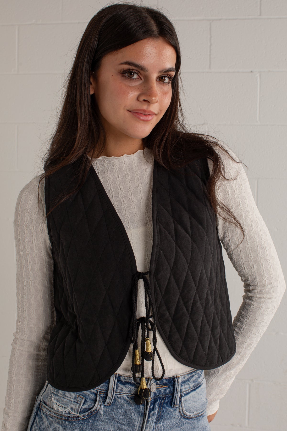 A woman wearing a black diamond-quilted sleeveless vest over a white long-sleeve top and blue jeans, standing against a white brick wall.