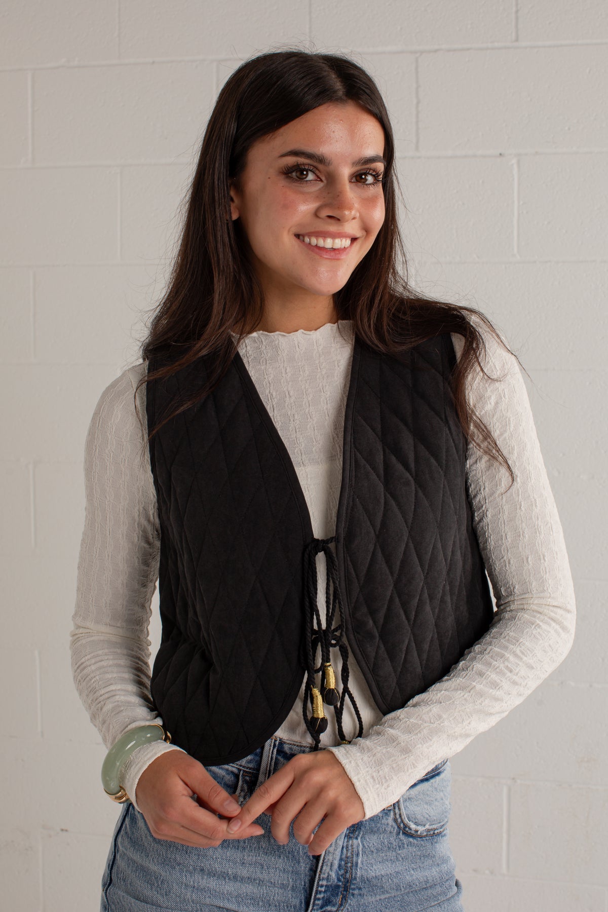 A woman wearing a black quilted sleeveless vest over a light gray long-sleeve top and blue jeans, standing against a white painted brick wall.