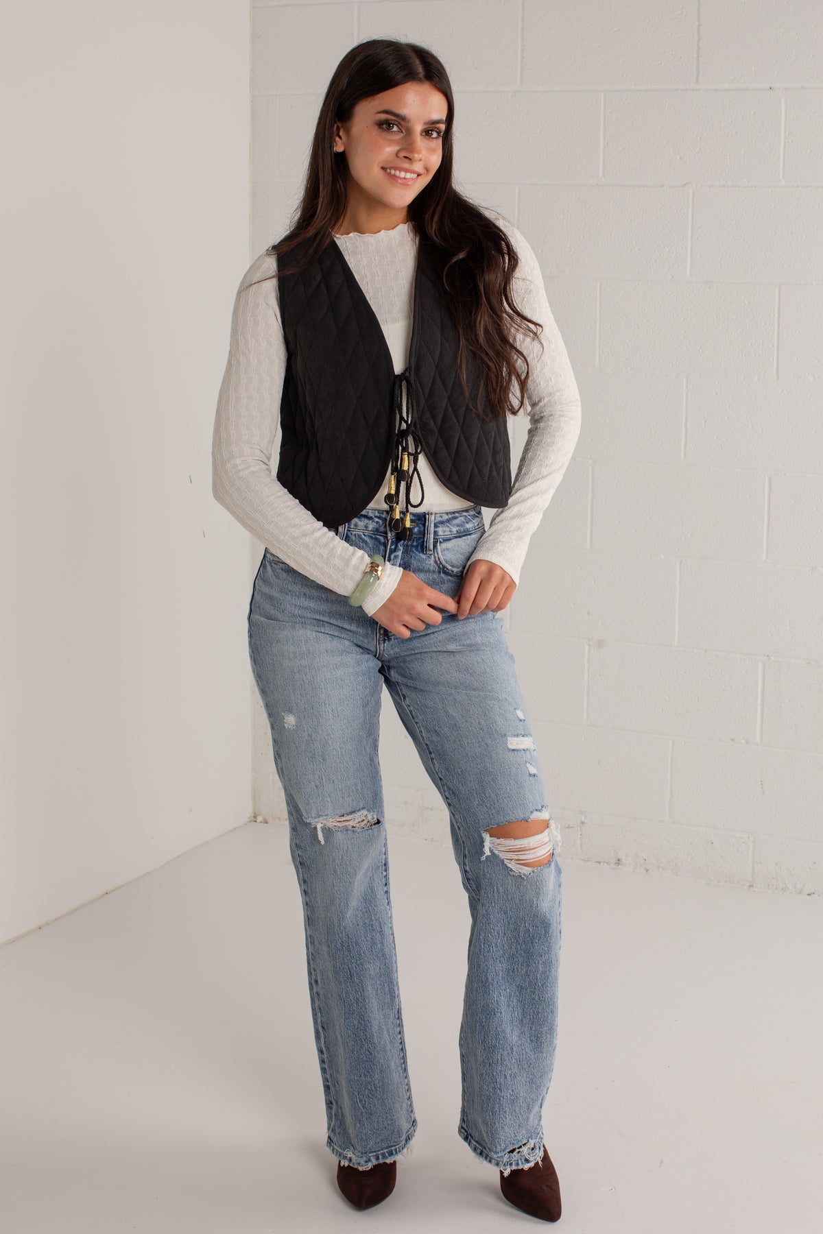 A woman modeling a black quilted vest over a white ribbed long-sleeve top, paired with light-wash distressed jeans and dark brown pointed-toe flats, standing against a white brick wall.