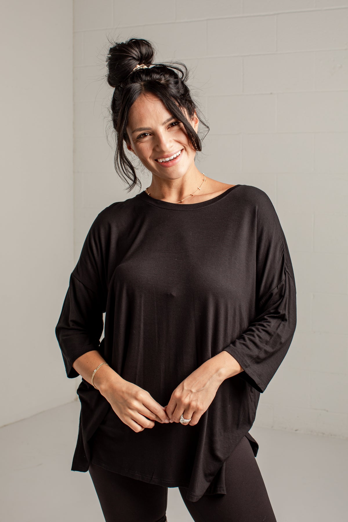 A woman with her hair in a bun wearing a loose-fitting black long-sleeve top and black leggings, standing against a light gray brick wall.