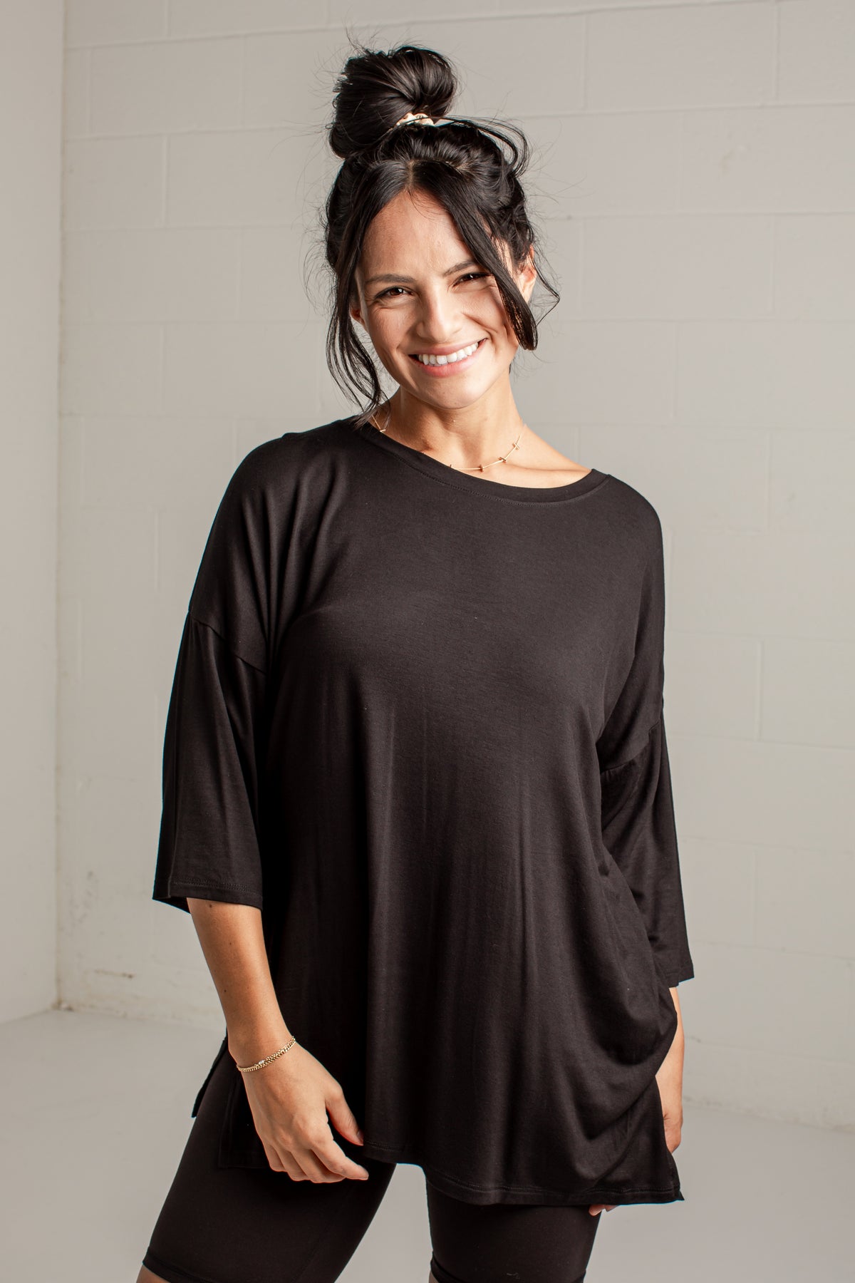 A woman with her hair in a bun wearing a loose-fitting black three-quarter sleeve top and black leggings, standing against a light gray brick wall.