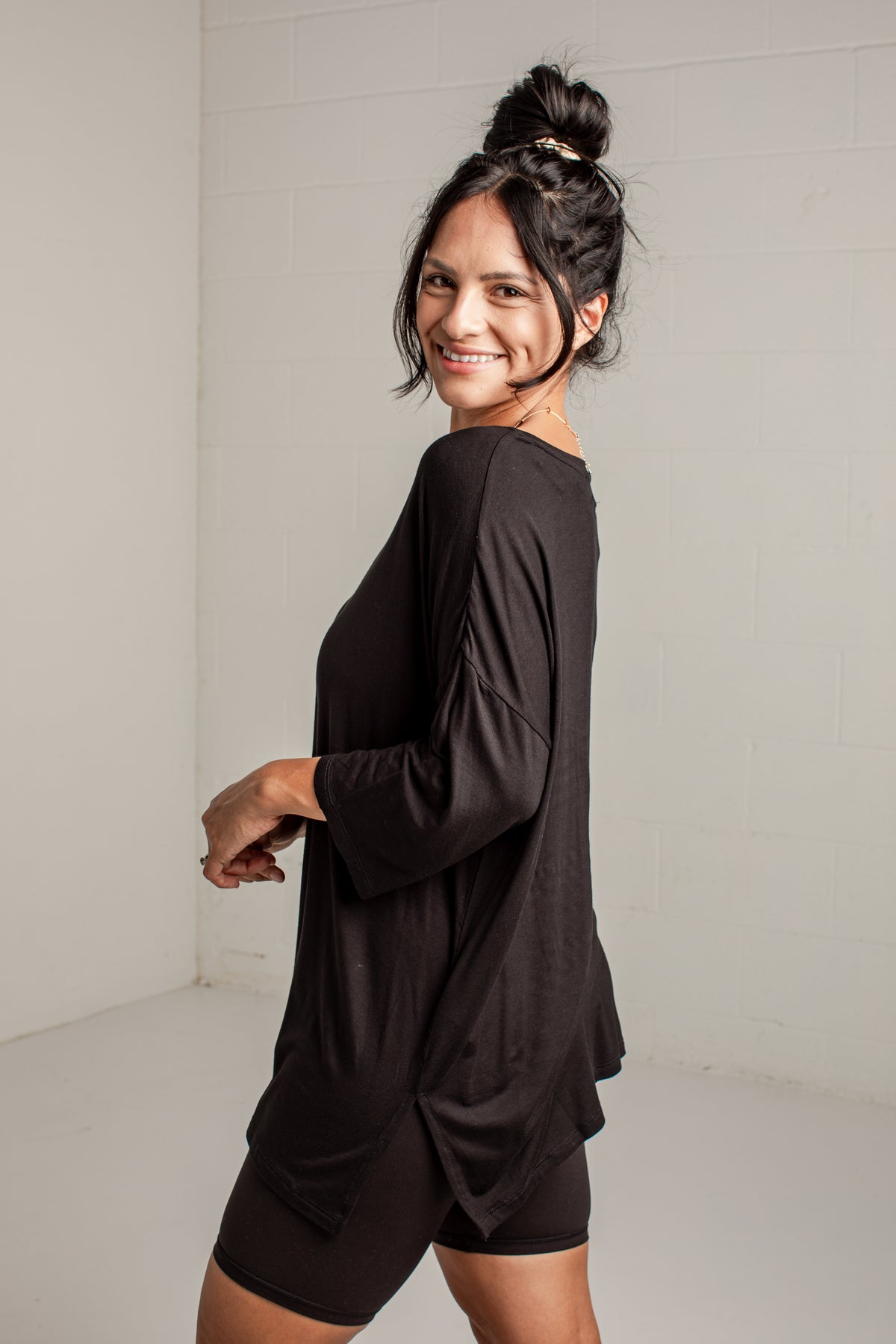 A woman wearing a loose black long-sleeve tunic top and matching black shorts, posing in a bright white studio setting.