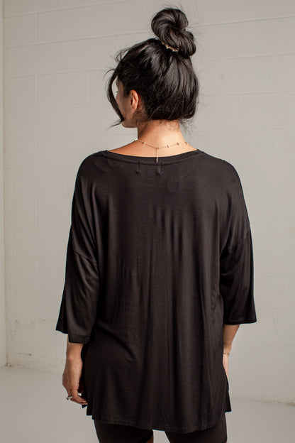 A woman viewed from behind wearing a loose black three-quarter sleeve top, standing against a light gray brick wall.
