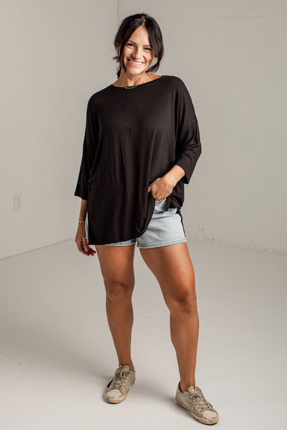 A woman modeling a loose-fitting black long-sleeve top with light denim shorts and beige sneakers, standing in a bright white studio.