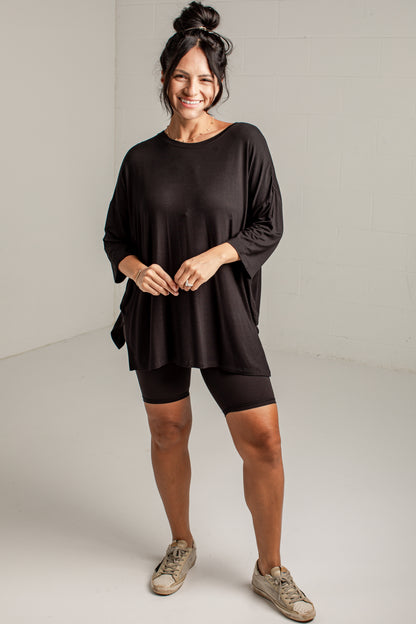 A woman modeling a black oversized three-quarter sleeve top with matching black biker shorts and beige sneakers, standing against a light gray studio wall.