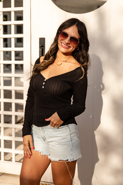 Woman wearing a black long-sleeve top and denim shorts, standing against a white wall.