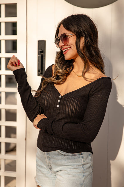 Woman wearing a black ribbed sweater and sunglasses against a white wall.