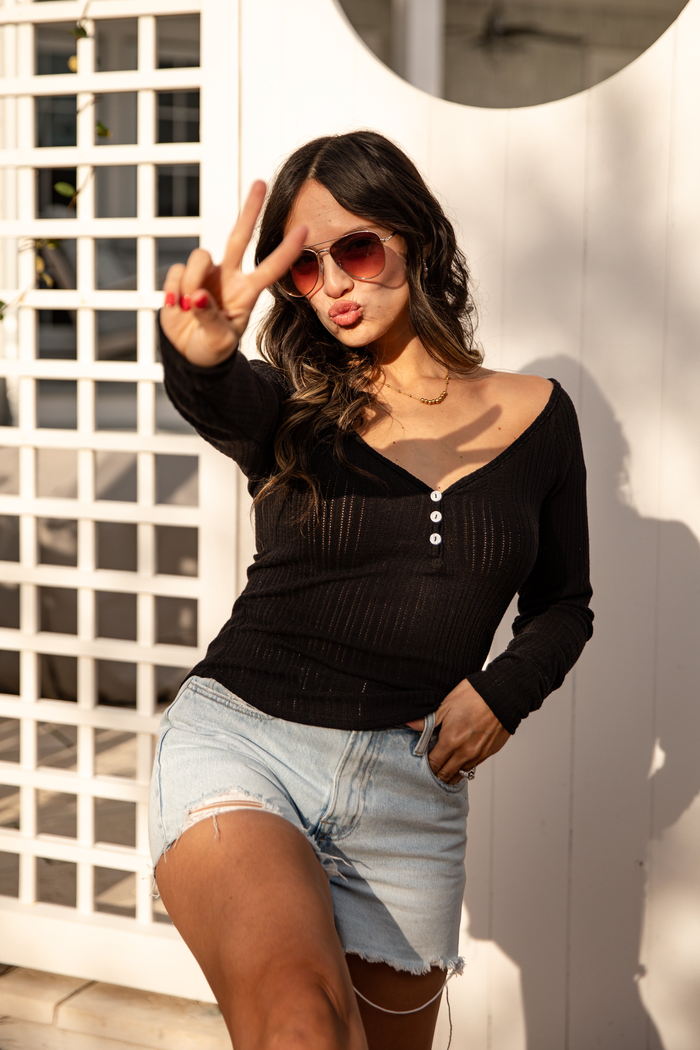 Woman wearing sunglasses and a black top with denim shorts, posing against a white wall.
